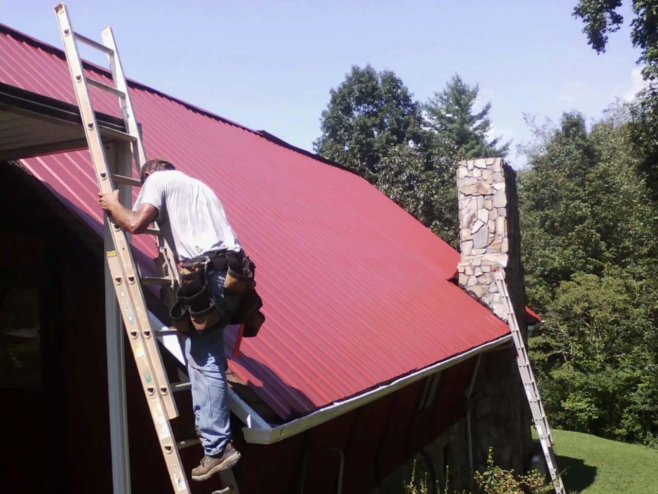 Roofer on a ladder beside a red metal roof and stone chimney — Soffit & Fascia Repair by Roofing Brothers in Hacienda Heights